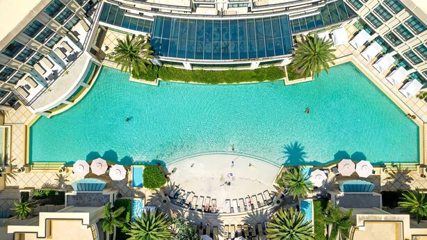 Une grande piscine aux eaux cristallines, entourée de chaises longues et de palmiers, avec un bâtiment moderne en arrière-plan.