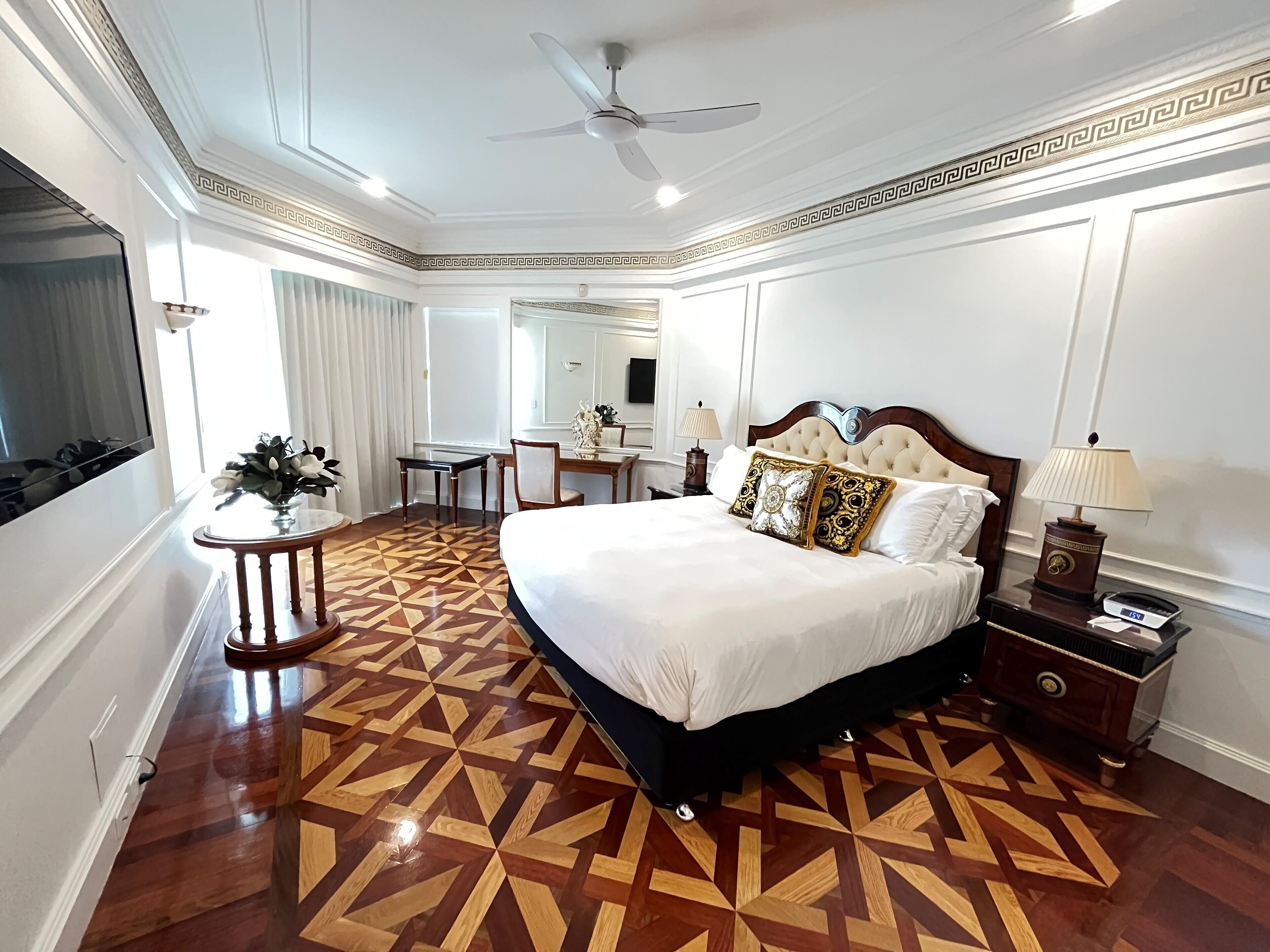 A hotel room with a large bed, a desk, a chair, a small table with a vase, a lamp, and a ceiling fan.
