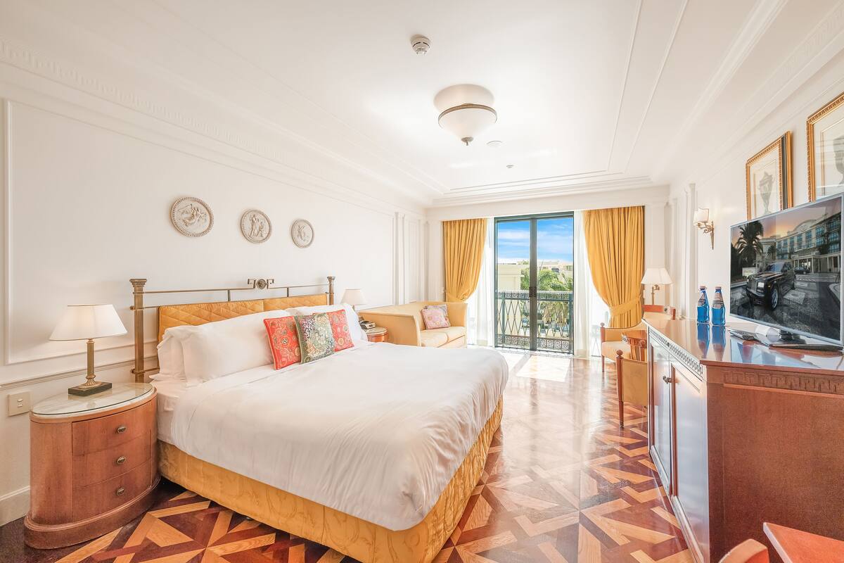 A hotel room with a large bed, a desk with a TV, and a balcony with curtains.