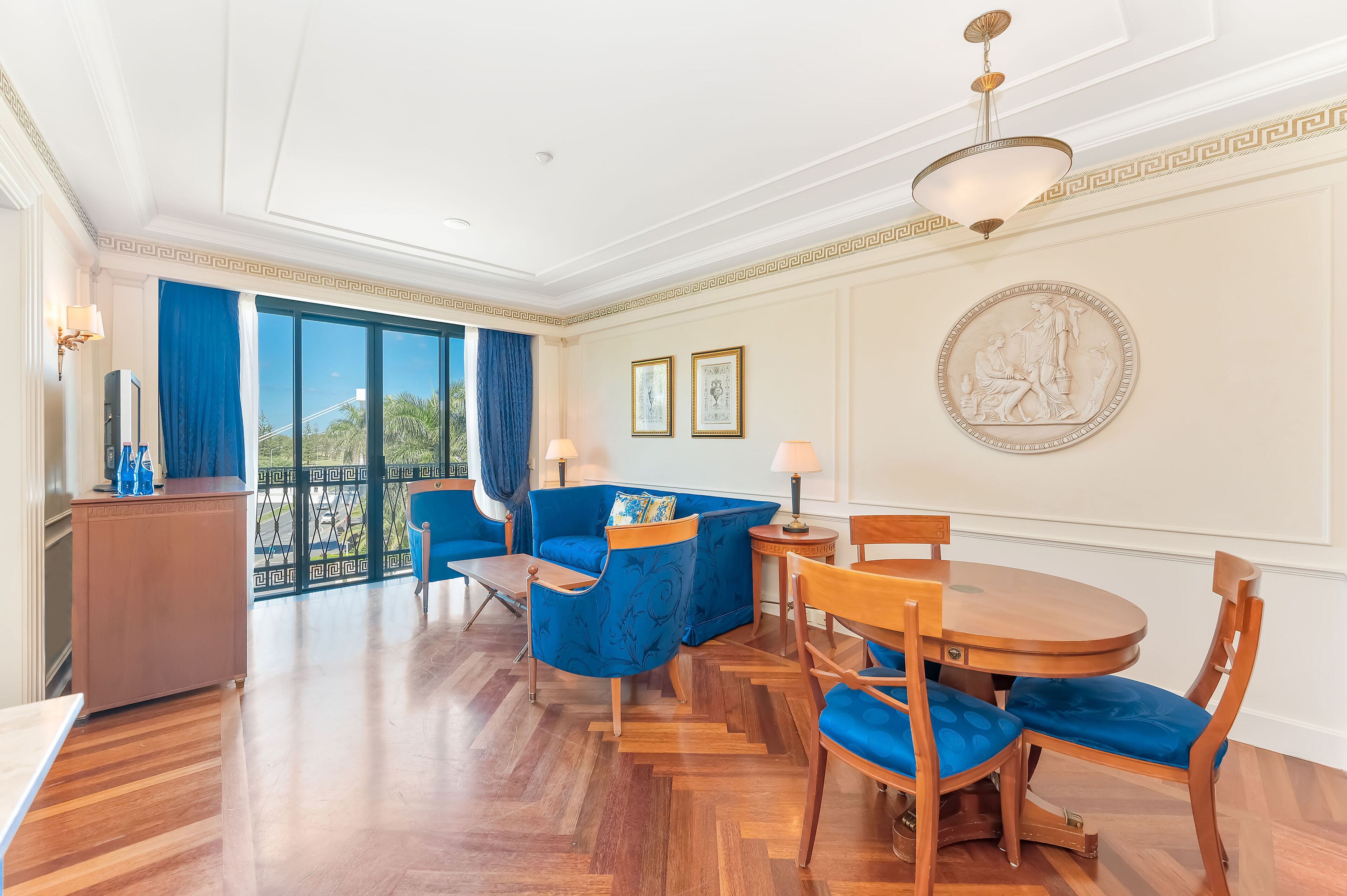 A spacious room with a balcony, a dining area with a round table and chairs, and a large window with curtains.