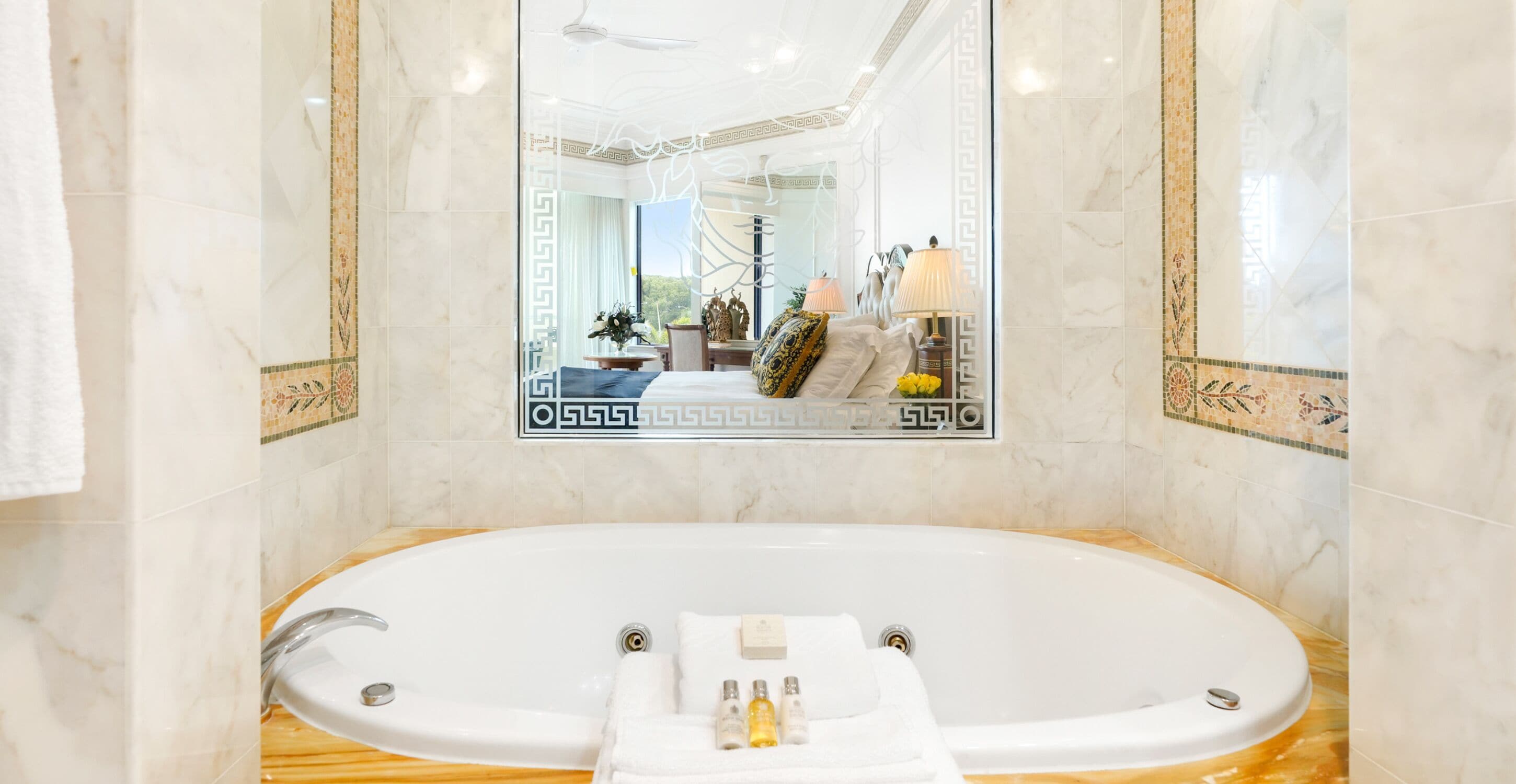 Un baño lujoso con una bañera grande, paredes de mármol y un espejo que refleja una habitación elegantemente decorada.