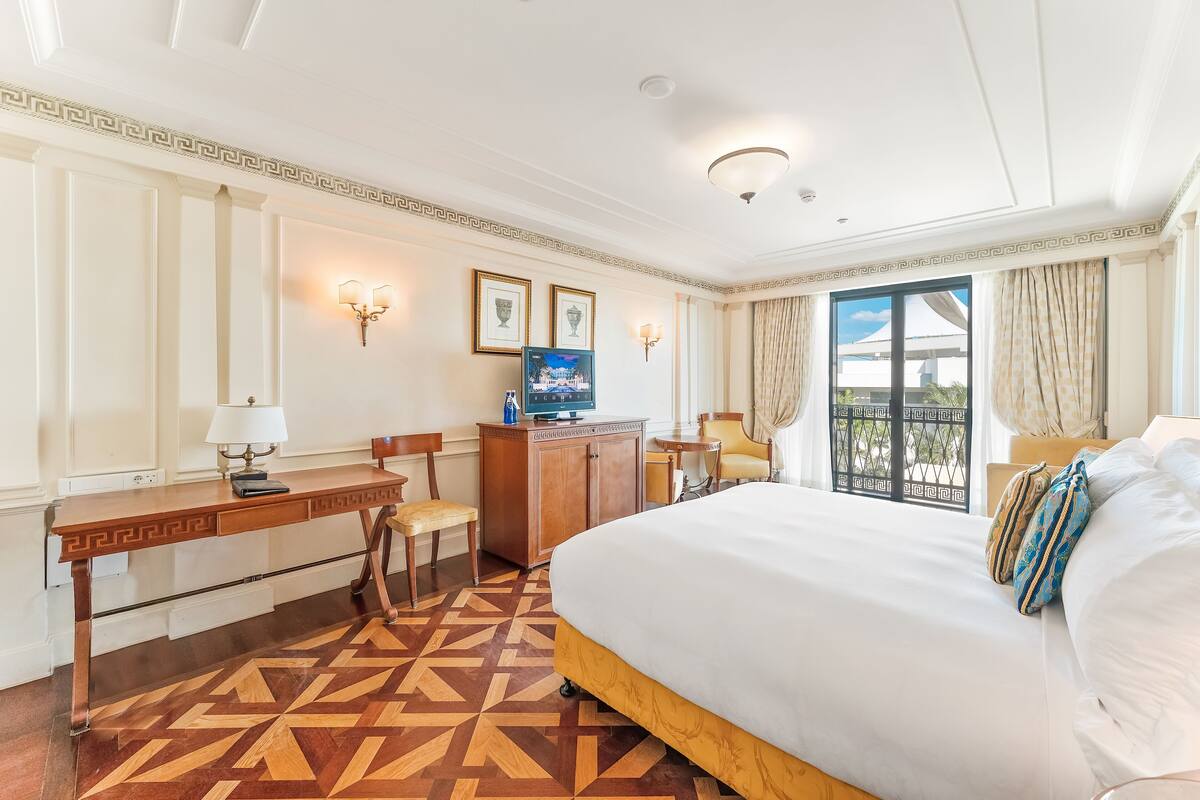 Habitación de hotel con cama, escritorio, silla, televisor y vistas a través de un ventanal grande.