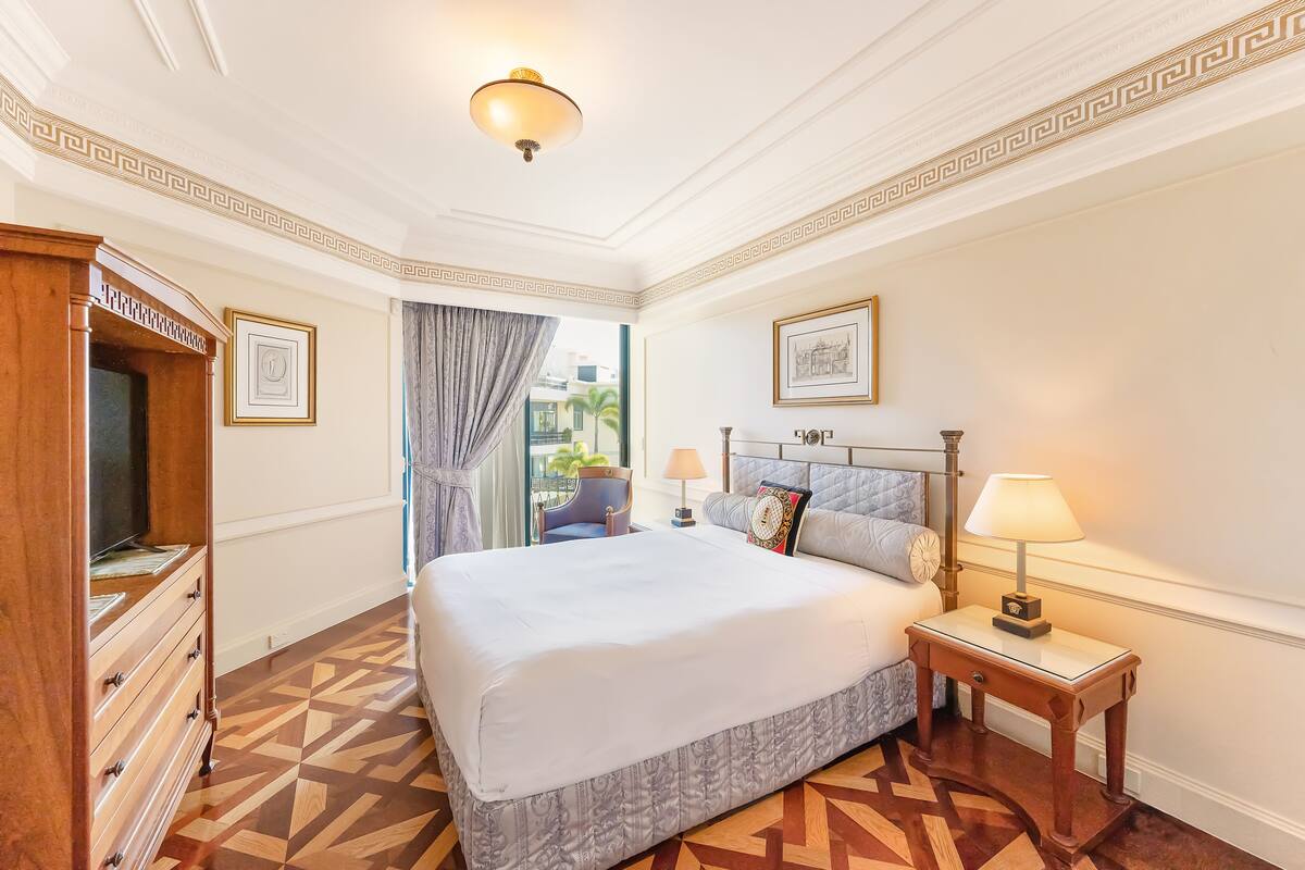 A hotel room with a large bed, a wooden dresser, a nightstand with a lamp, and a view of the outdoors.