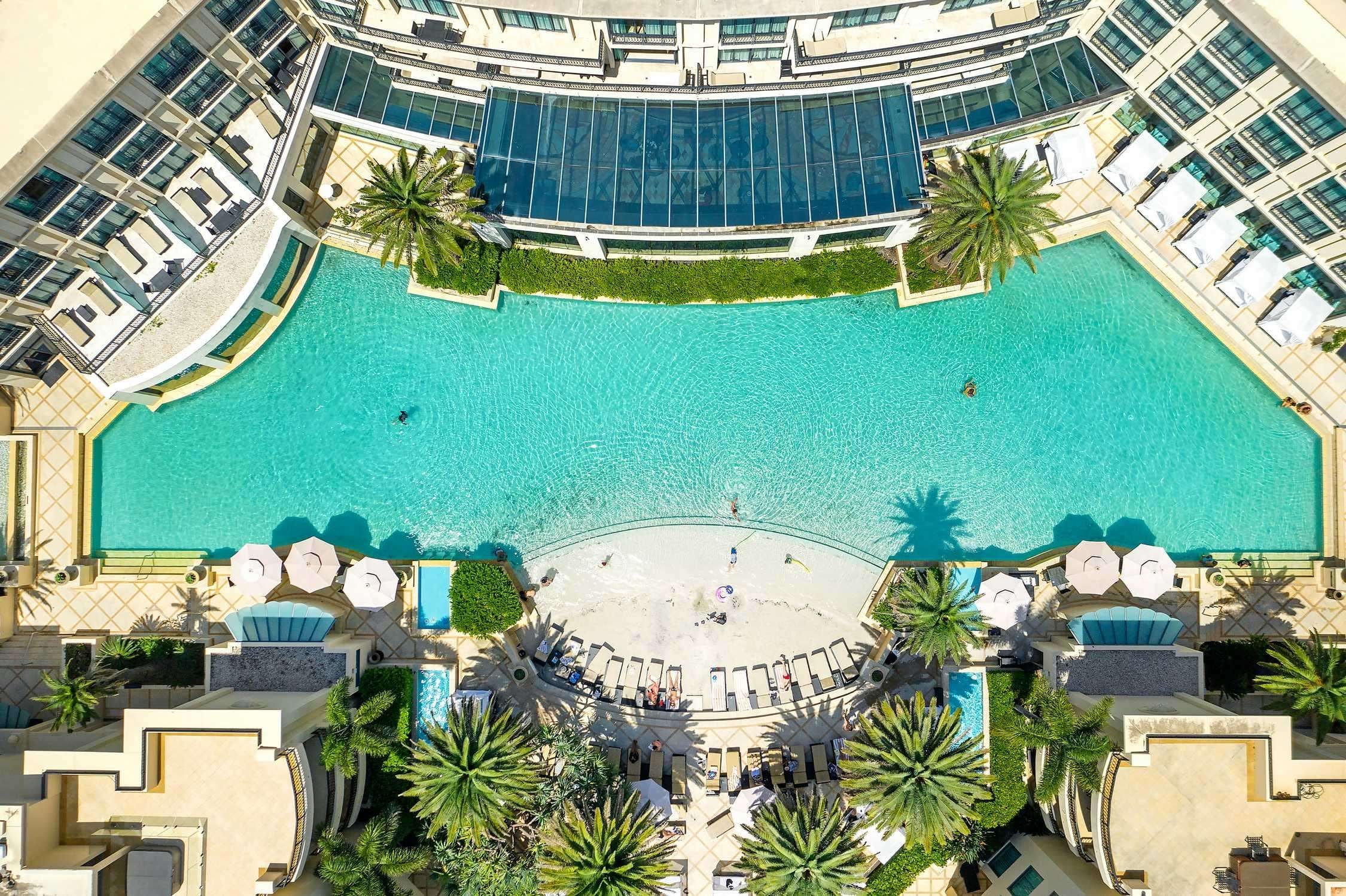Una piscina amplia y cristalina con terraza solárium, rodeada de sillones de descanso y palmeras.
