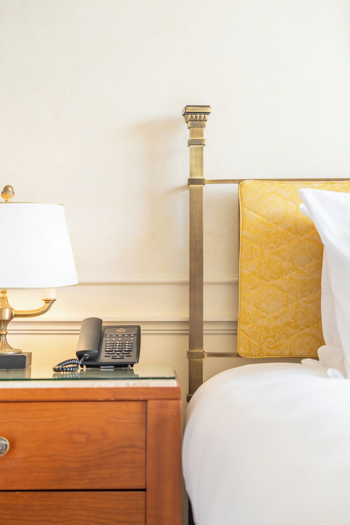 A hotel room with a wooden nightstand, a telephone, a lamp, and a bed with a patterned headboard.