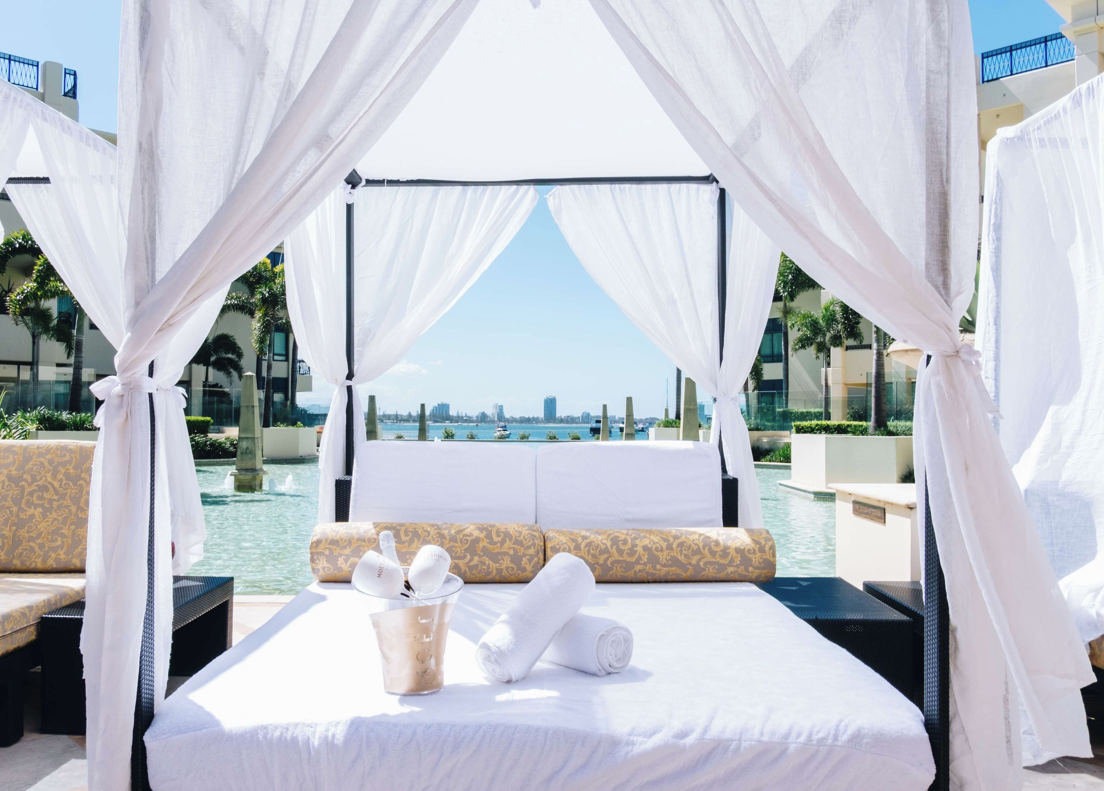 Een bed met witte lakens, een glas water en een handdoek, met op de achtergrond een stadszicht en een zwembad.