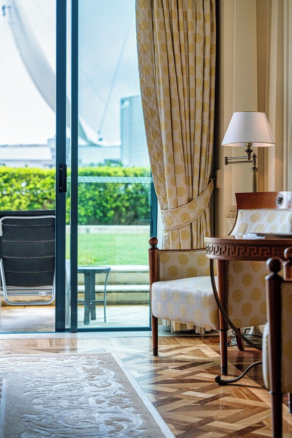 A hotel room with a large window, a chair, a bedside table with a lamp, and a patterned rug.