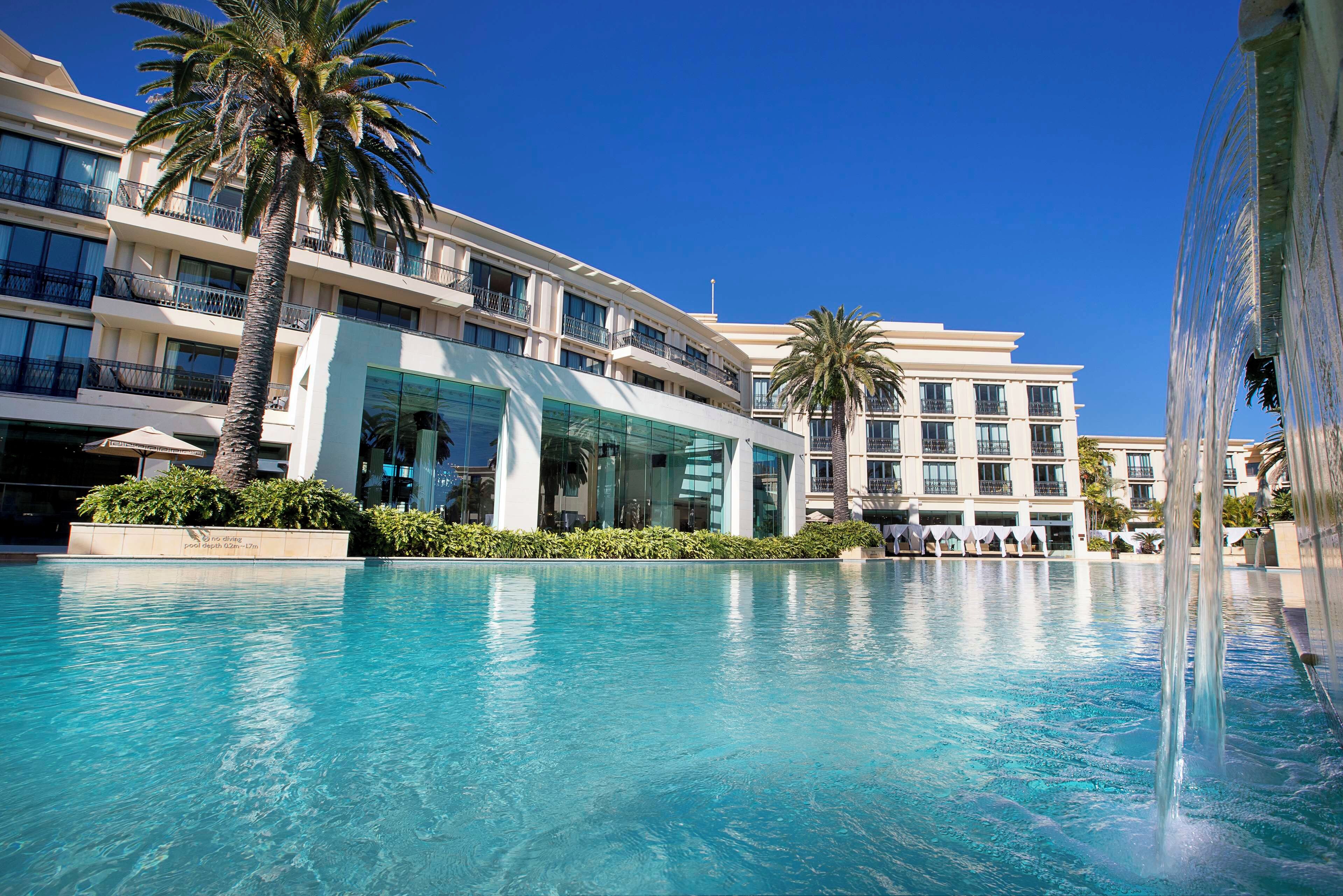 A hotel with a swimming pool, palm trees, and a clear blue sky.