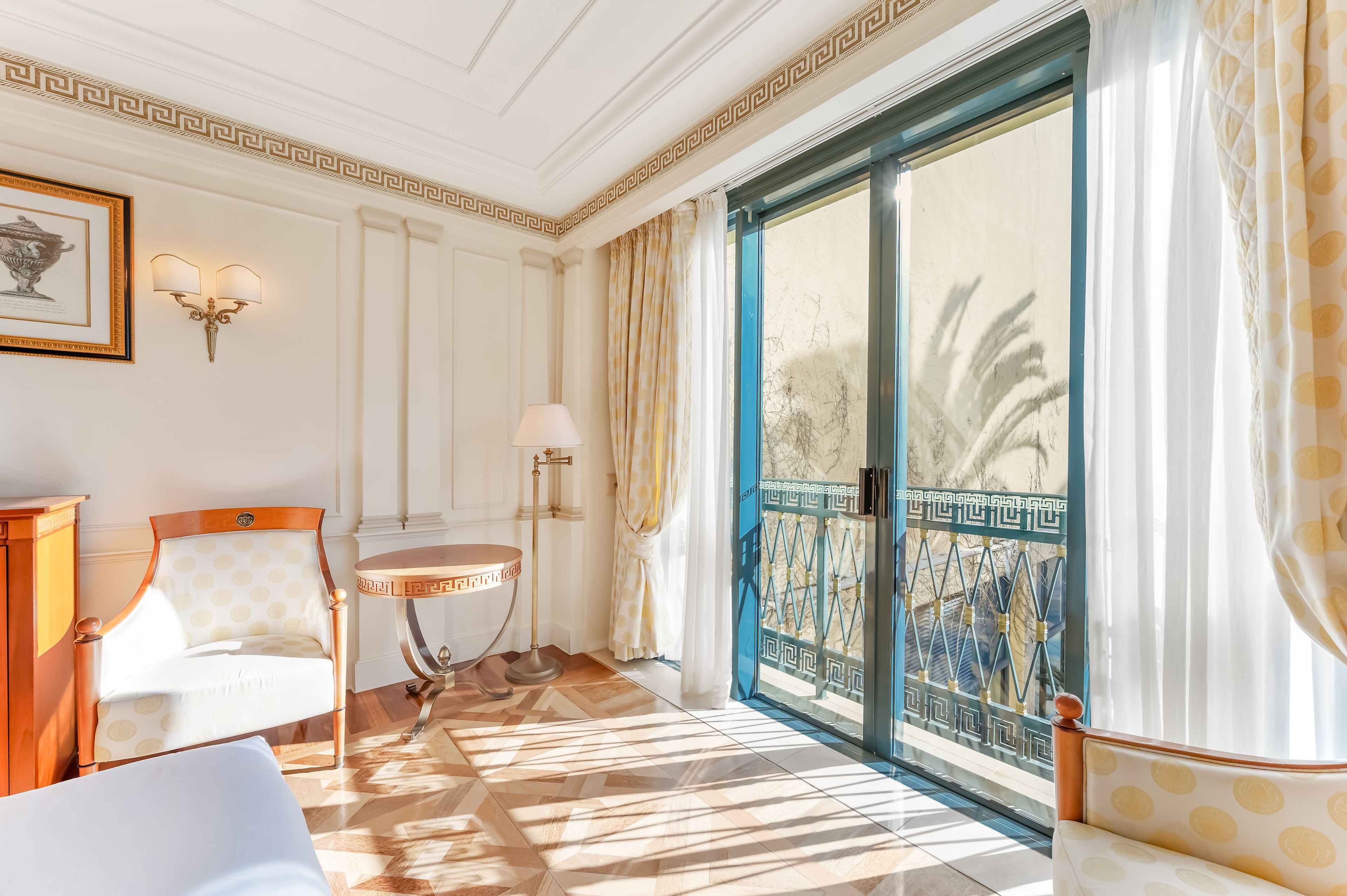 A room with a patterned chair, a round table, a wall-mounted lamp, and a large window with a view of palm trees.