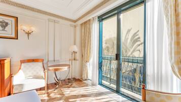 A room with a patterned chair, a round table, a wall-mounted lamp, and a large window with a view of palm trees.