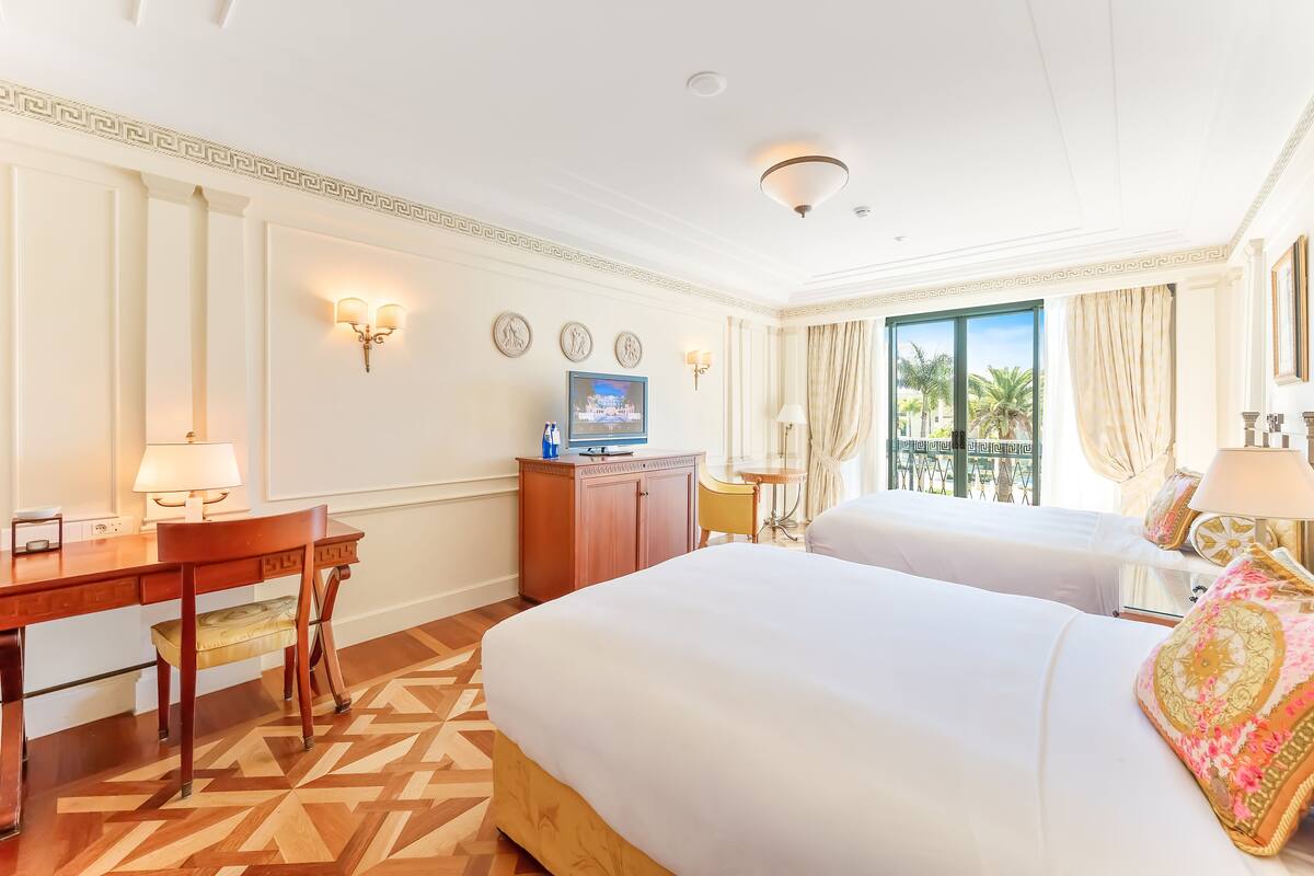 A hotel room with two beds, a desk, a TV, and a balcony with curtains.