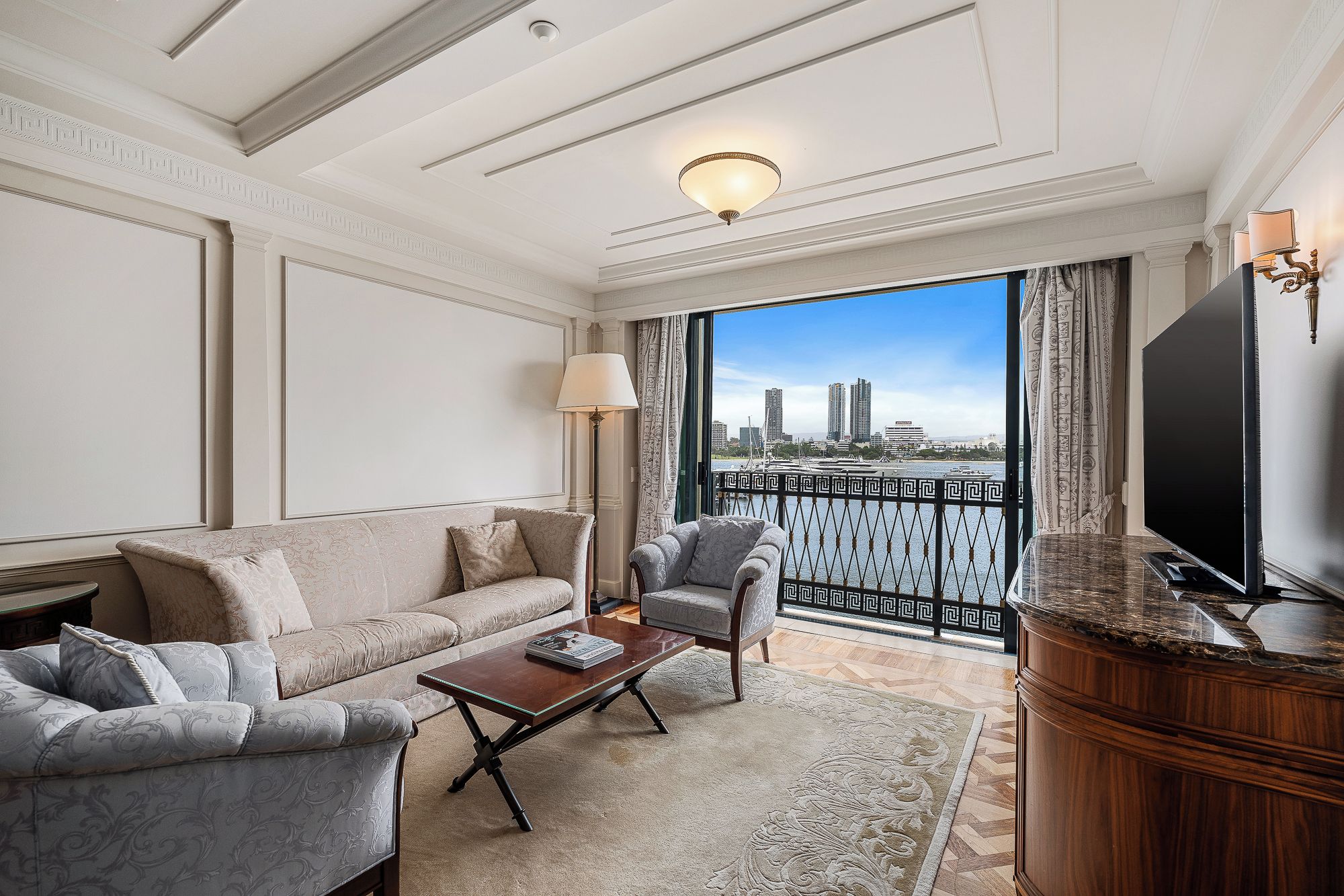 Habitación de hotel con sofá, sillón, mesa de centro, televisor y balcón con vistas a edificios y al agua.