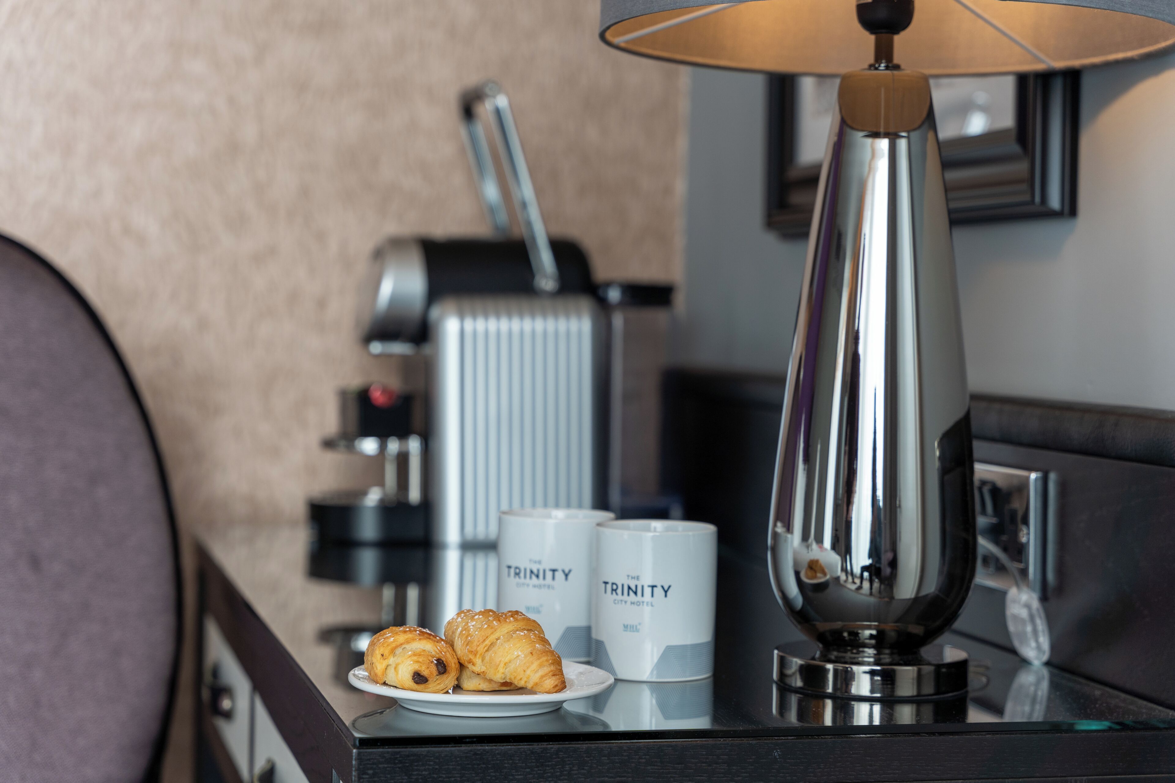 A hotel room with a coffee machine, two coffee cups labeled 'The Trinity City Hotel,' and two croissants on a plate.