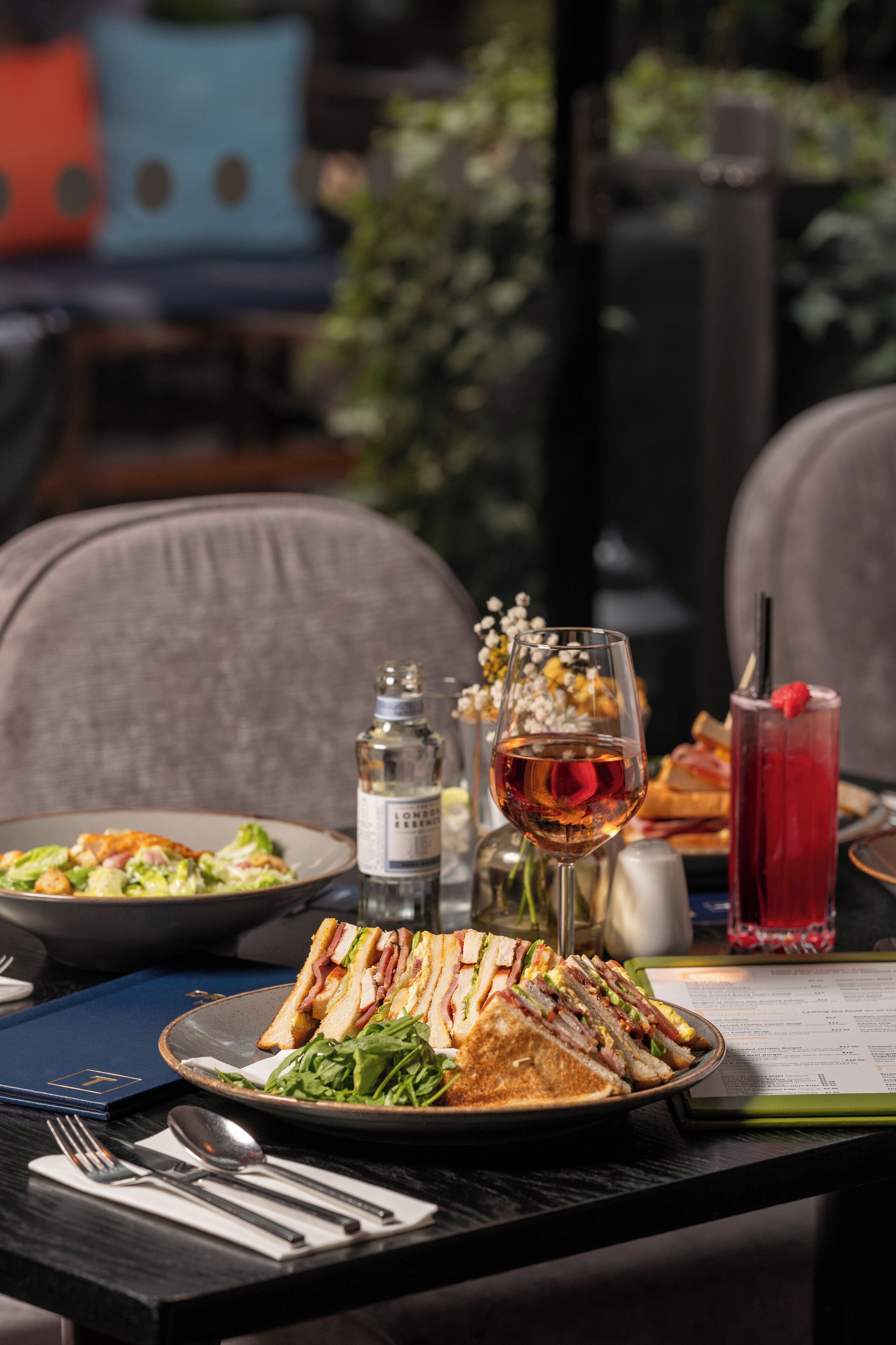 A dining table with a plate of sandwiches, a glass of wine, a red drink, and a menu.