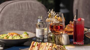A dining table with a plate of sandwiches, a glass of wine, a red drink, and a menu.