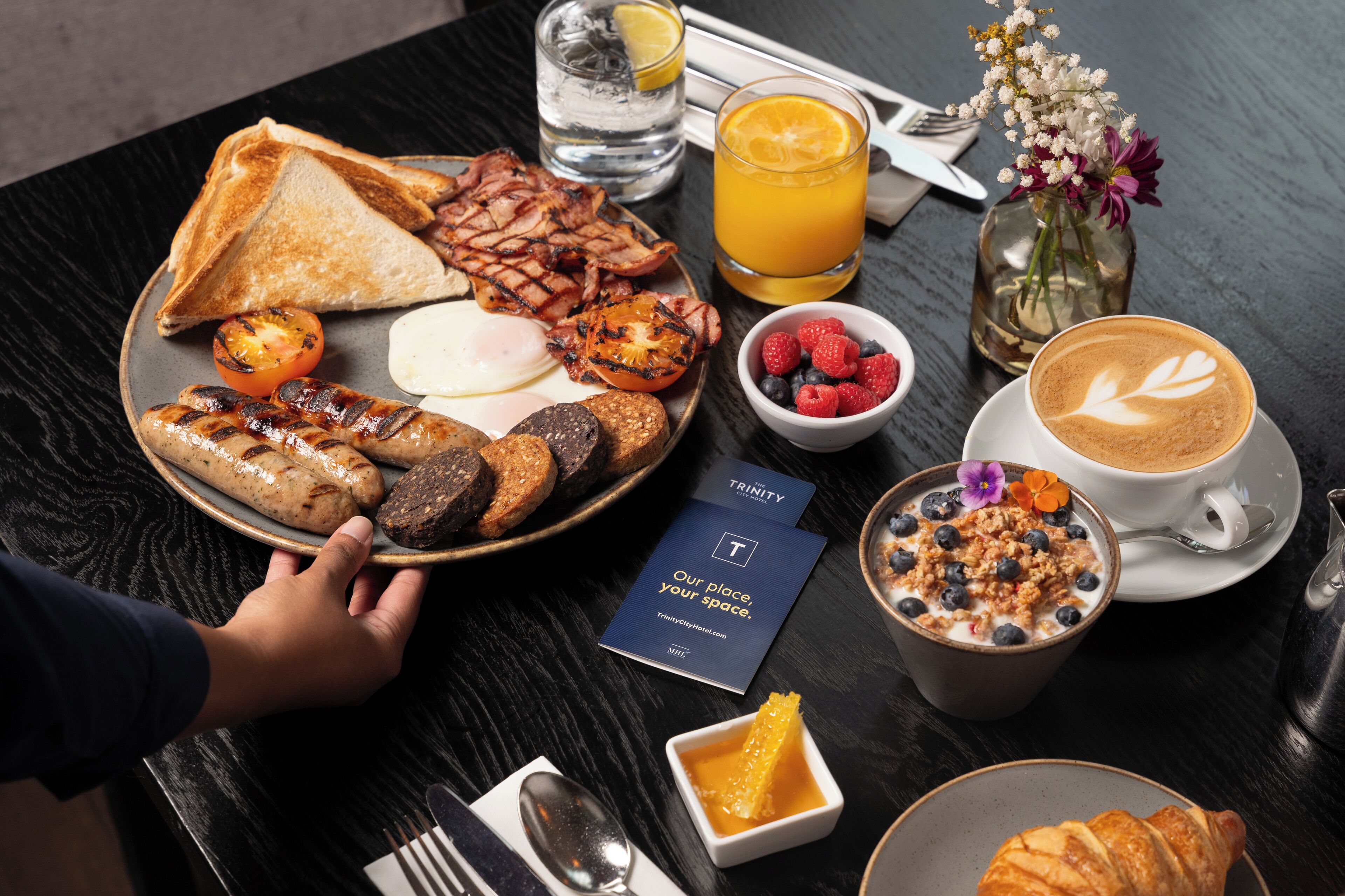 A breakfast plate with toast, bacon, eggs, and sausages, a glass of orange juice, a bowl of berries, a cup of coffee with latte art, and a croissant.