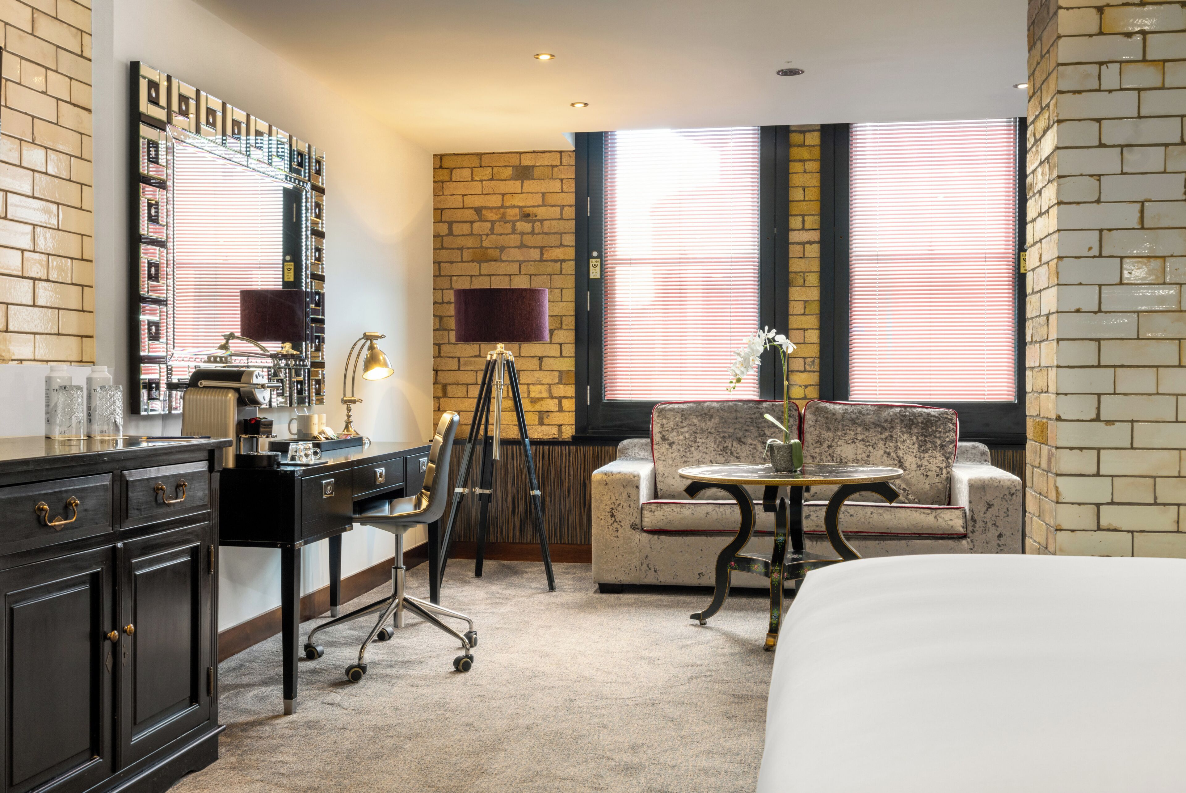A hotel room with a bed, a desk with a lamp, a chair, a small table, a sofa, a mirror, and a window with blinds.
