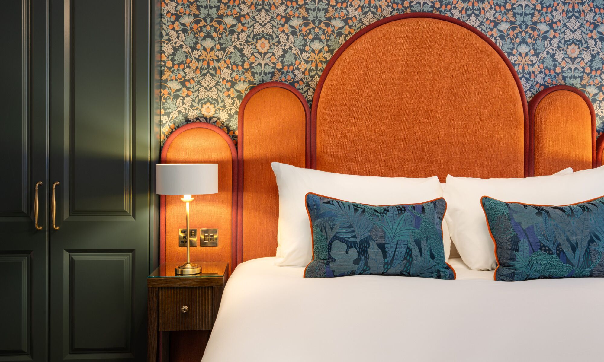 A hotel room with a large bed, two pillows, a nightstand with a lamp, and a wall with a floral pattern.