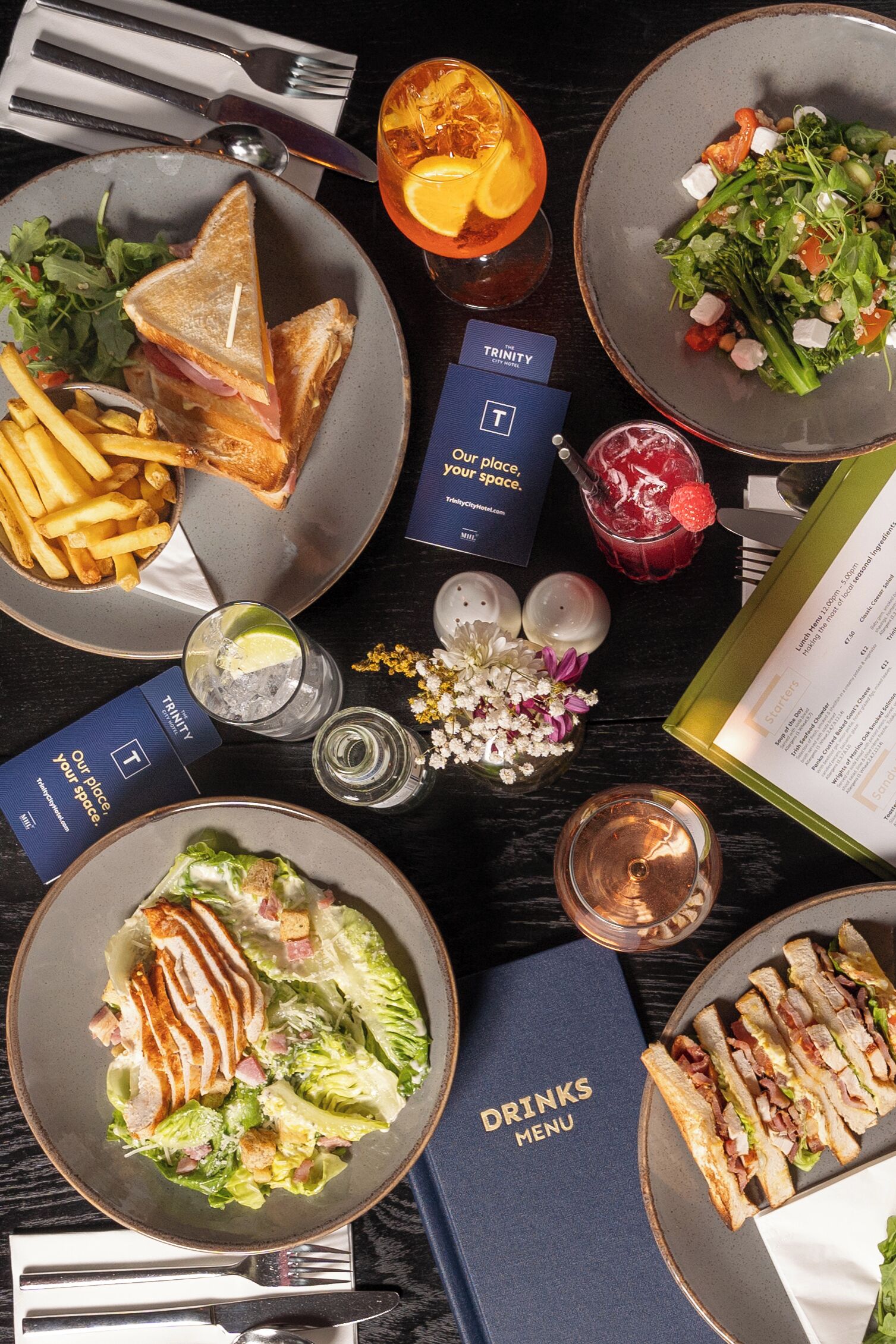 Una mesa dispuesta con diversos platos, incluyendo una ensalada, sándwiches y una bebida, junto con un menú y algunos adornos.