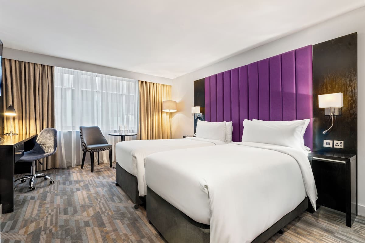 Hotellihuone, jossa on kaksi sänkyä, työpöytä, tuoli ja suuri ikkuna, jossa on verhot.