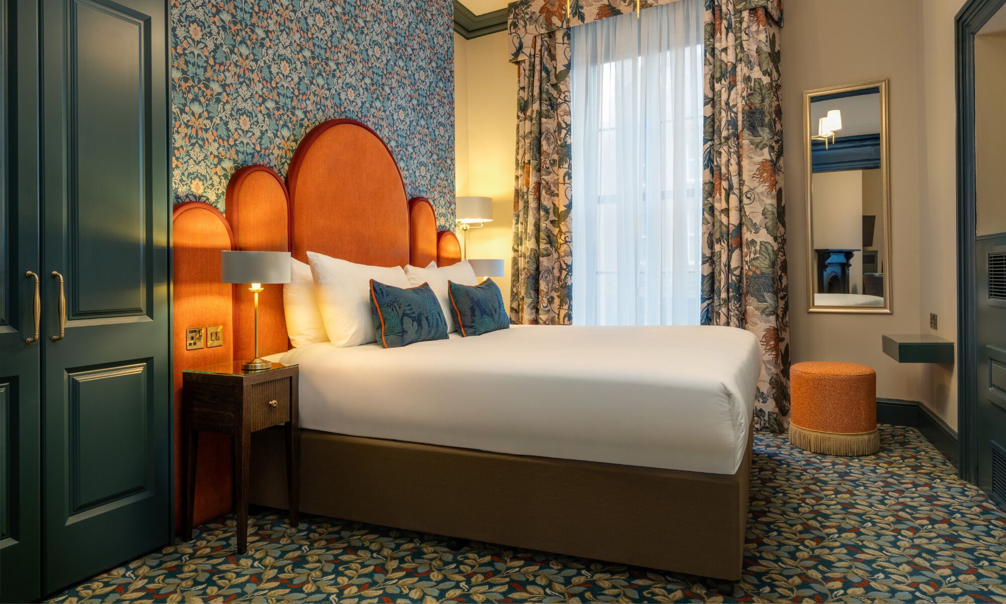 A hotel room with a large bed, a nightstand with a lamp, a small stool, and a mirror on the wall.