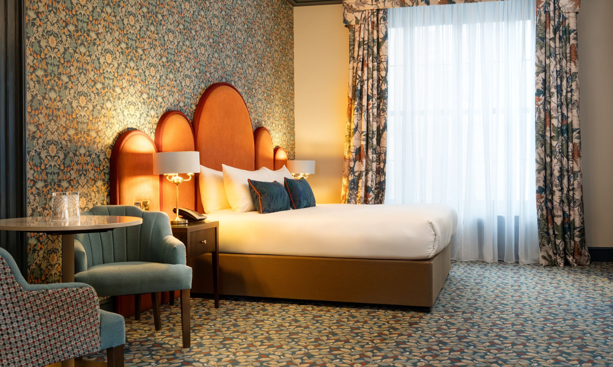 Habitación de hotel con una cama grande, dos lámparas de noche, una mesita con un teléfono, una silla y una alfombra estampada.