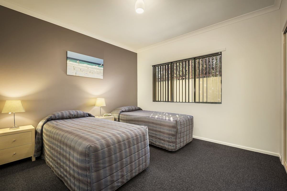 Une chambre d’hôtel avec deux lits, une table de chevet avec une lampe, une fenêtre avec des stores et un tableau au mur.
