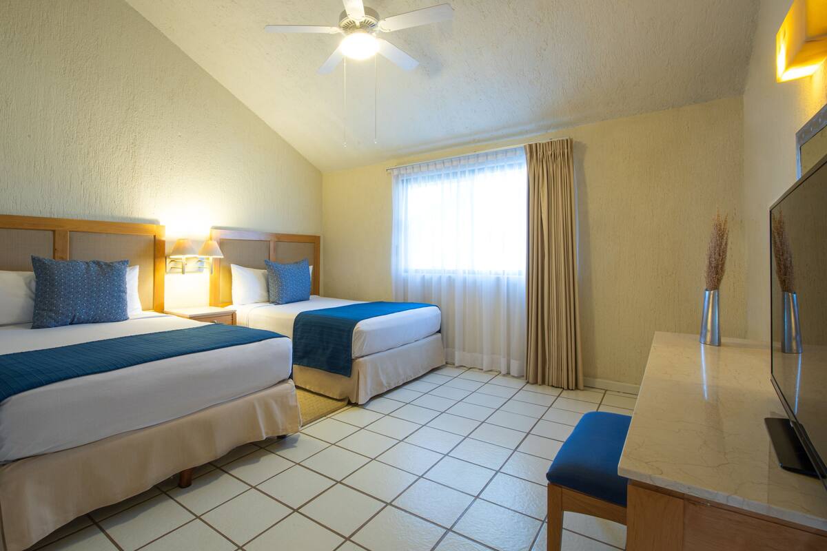 A hotel room with two beds, a ceiling fan, a window with curtains, a bedside table with lamps, and a mirror.