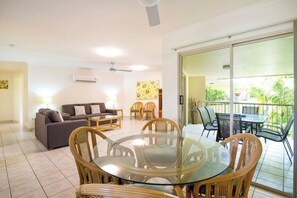 A living room with a sofa, a glass-top dining table, and chairs, a balcony with outdoor furniture, and a sliding glass door. - Port Douglas Sands Resort (Port Douglas)