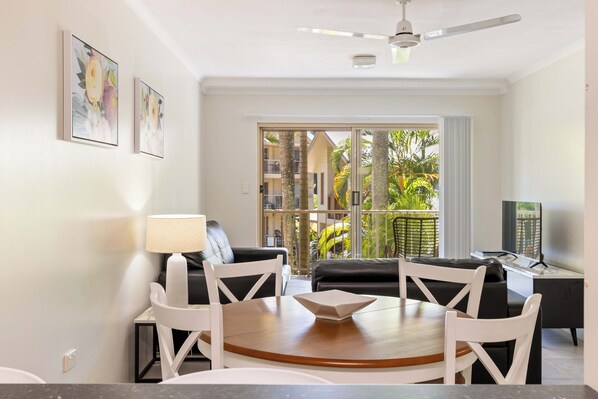 A modern living room with a dining area, a ceiling fan, and a balcony with a view of palm trees.
