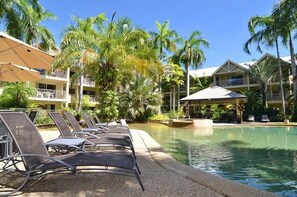 A poolside area with lounge chairs, palm trees, and a building with balconies. - Port Douglas Sands Resort (Port Douglas)