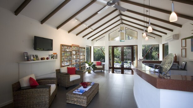 Uma sala de estar ampla, com móveis de vime, uma televisão de tela plana, uma estante de livros e uma grande janela com vista para o exterior.
