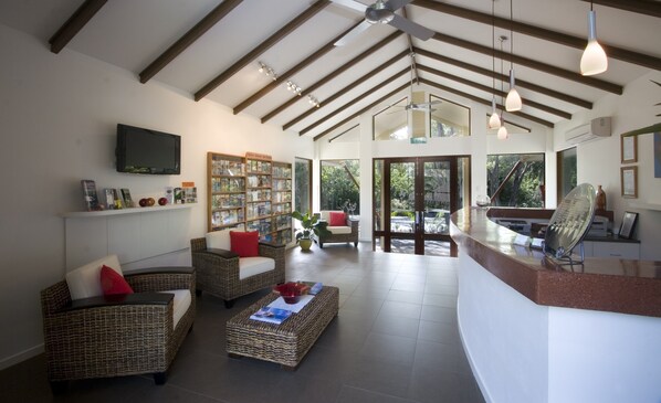 A spacious living area with wicker furniture, a flat-screen TV, a bookshelf, and a large window offering a view of the outdoors.