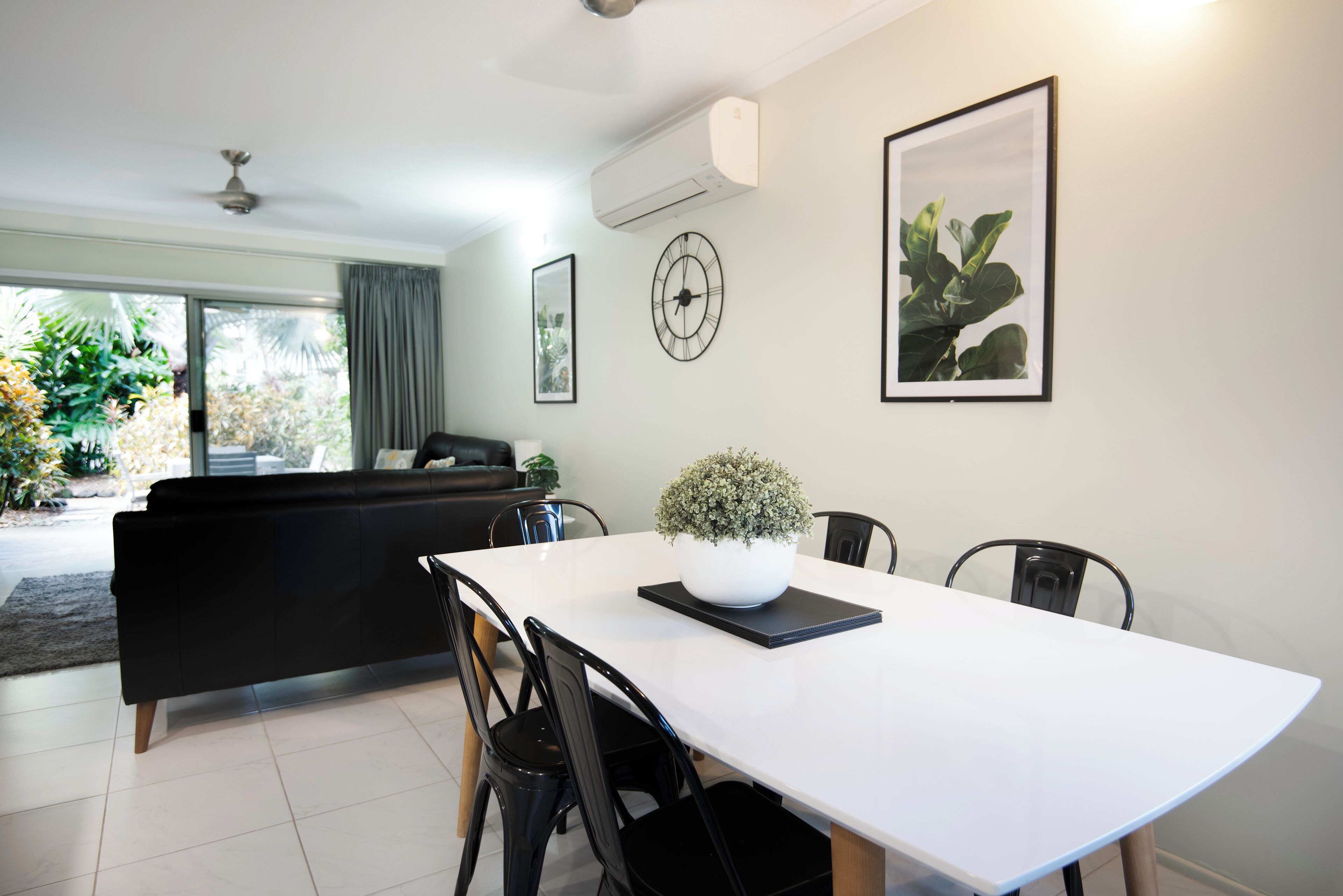 A modern living room with a white dining table, black chairs, a black sofa, and a potted plant.