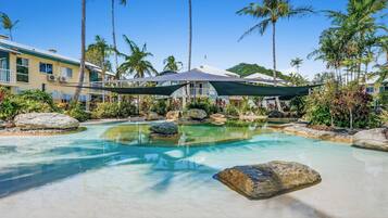 A resort pool with clear water, surrounded by rocks and palm trees, with a building in the background.