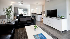 A modern living room with a black leather sofa, a white coffee table with a book, and a television on a white cabinet. - Marlin Cove Resort (Trinity Beach)