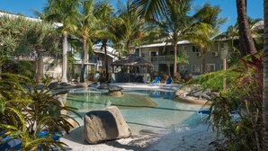 A resort with a pool surrounded by palm trees and rocks, a thatched-roof structure, and a building with balconies. - Marlin Cove Resort (Trinity Beach)