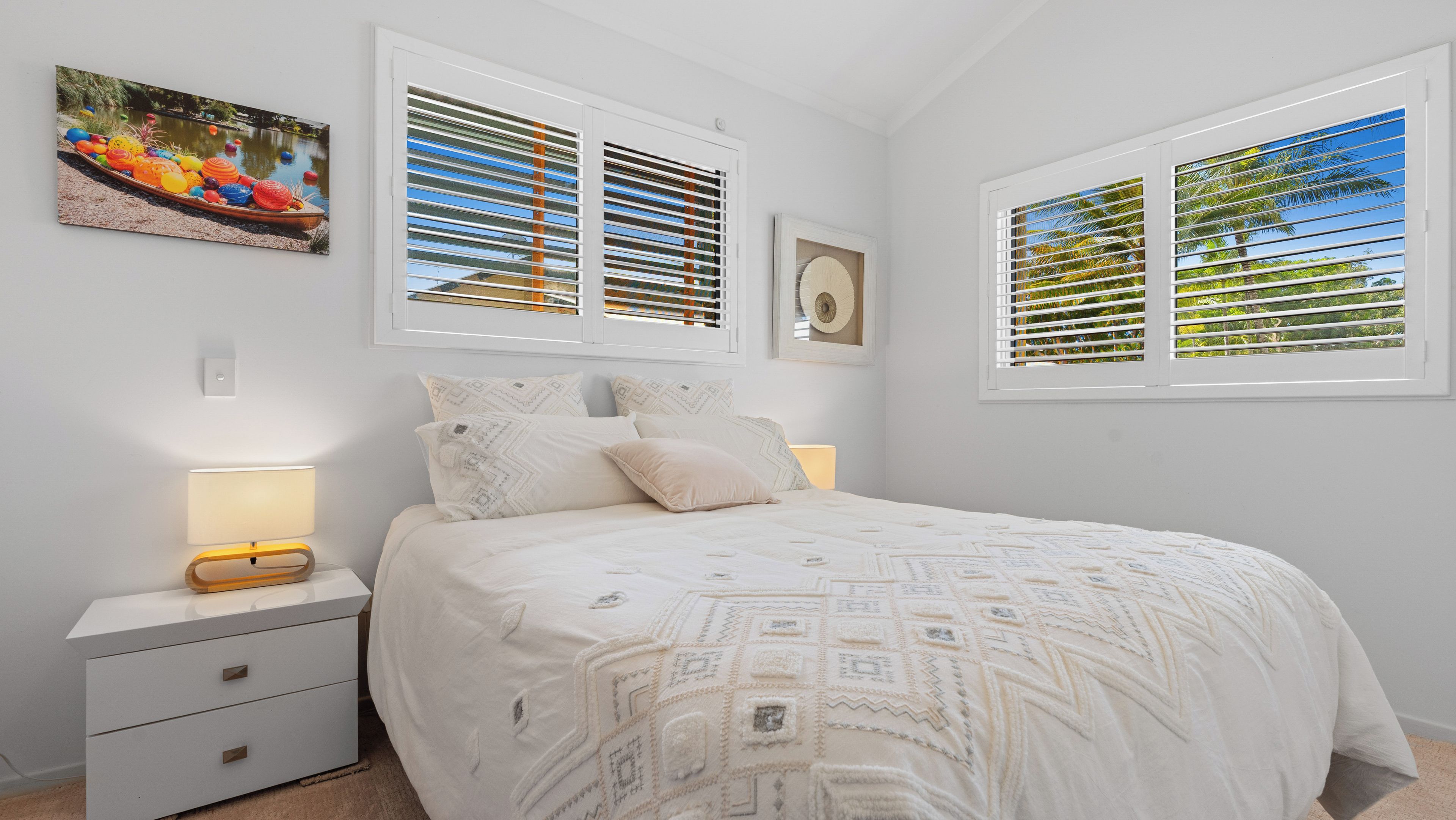Une chambre à coucher avec un lit, des tables de chevet, une lampe et deux fenêtres équipées de stores.