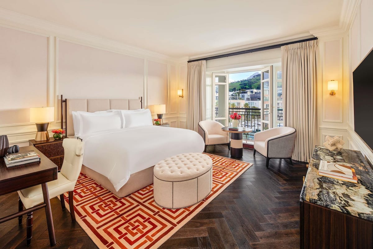 Habitación de hotel con una cama grande, un área de estar con una mesa y sillas, vista de la ciudad a través de un ventanal amplio y un televisor a la derecha.