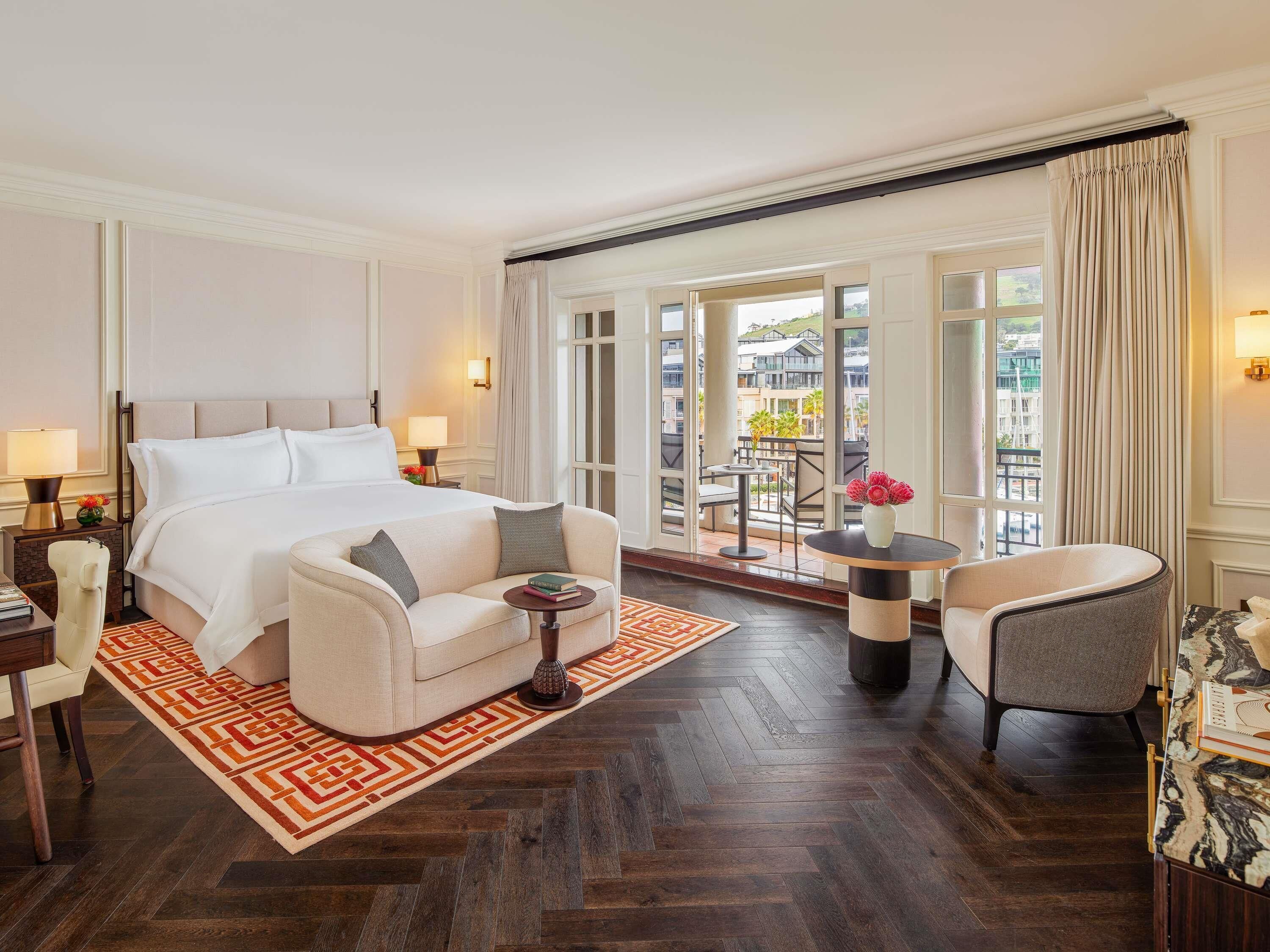 A spacious hotel room with a large bed, a sitting area with a sofa and armchairs, a small table with a vase, and a balcony with a view of the city.