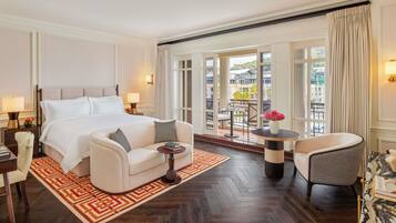 A spacious hotel room with a large bed, a sitting area with a sofa and armchairs, a small table with a vase, and a balcony with a view of the city.