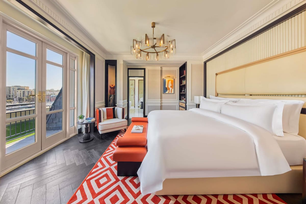 Habitación de hotel con una cama grande, un sofá, una araña y vistas de la ciudad, además de una puerta que da a otra habitación.