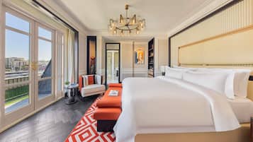A hotel room with a large bed, a sofa, a chandelier, a view of the city, and a door leading to another room.