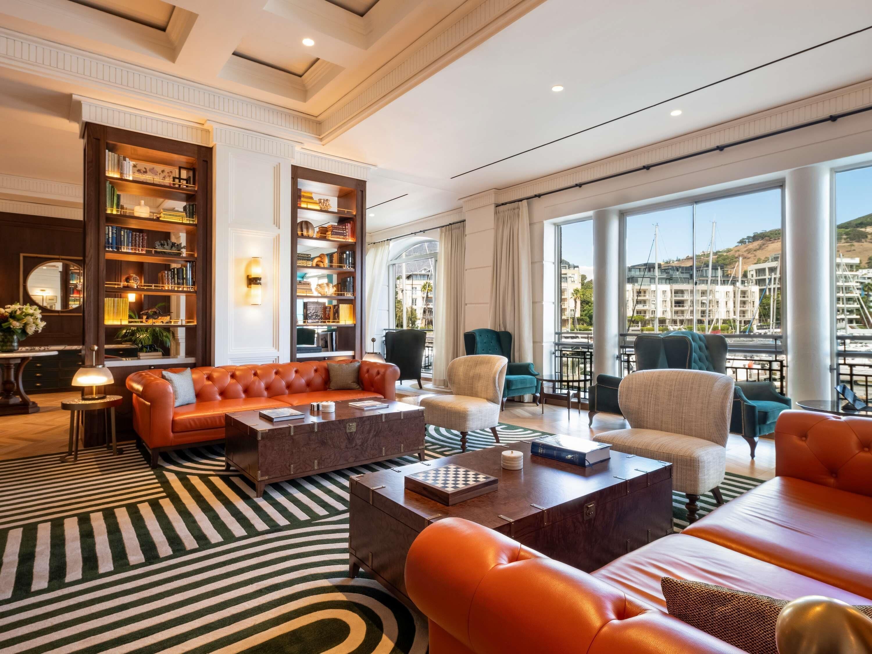 A spacious living room with a large sofa, multiple armchairs, a coffee table, and a fireplace. There are bookshelves filled with books and decorative items. The room has large windows offering a view of buildings outside.