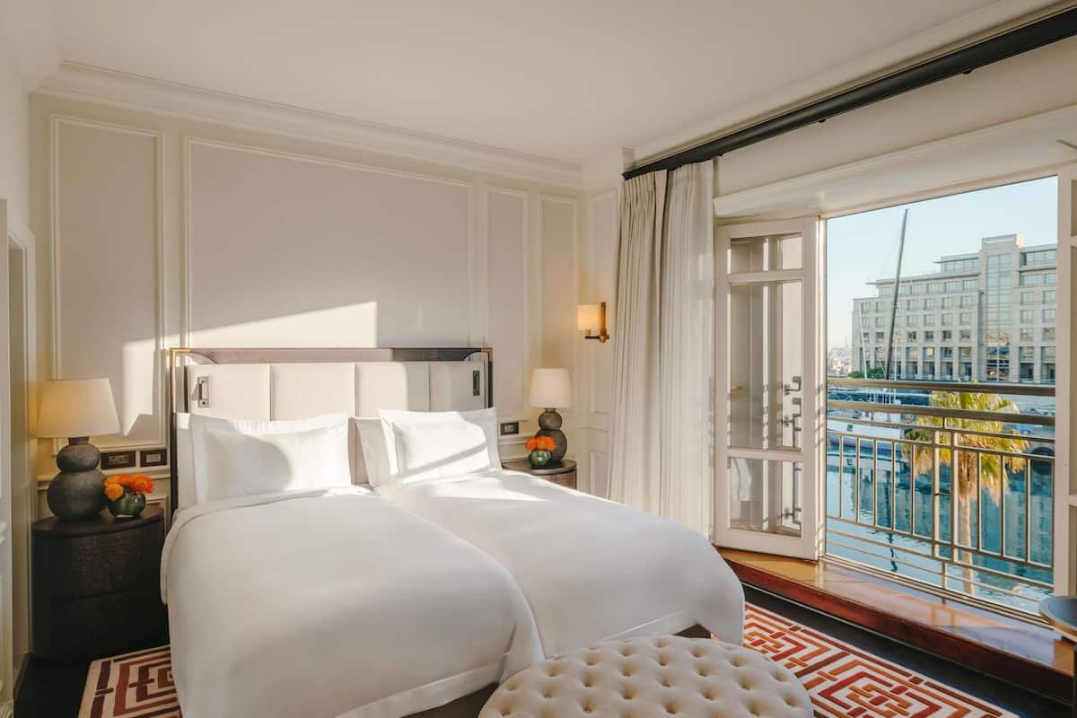 Habitación de hotel con una cama grande, un otomanillo redondo, una mesita de noche con lámpara y vistas a un paisaje urbano desde un balcón.