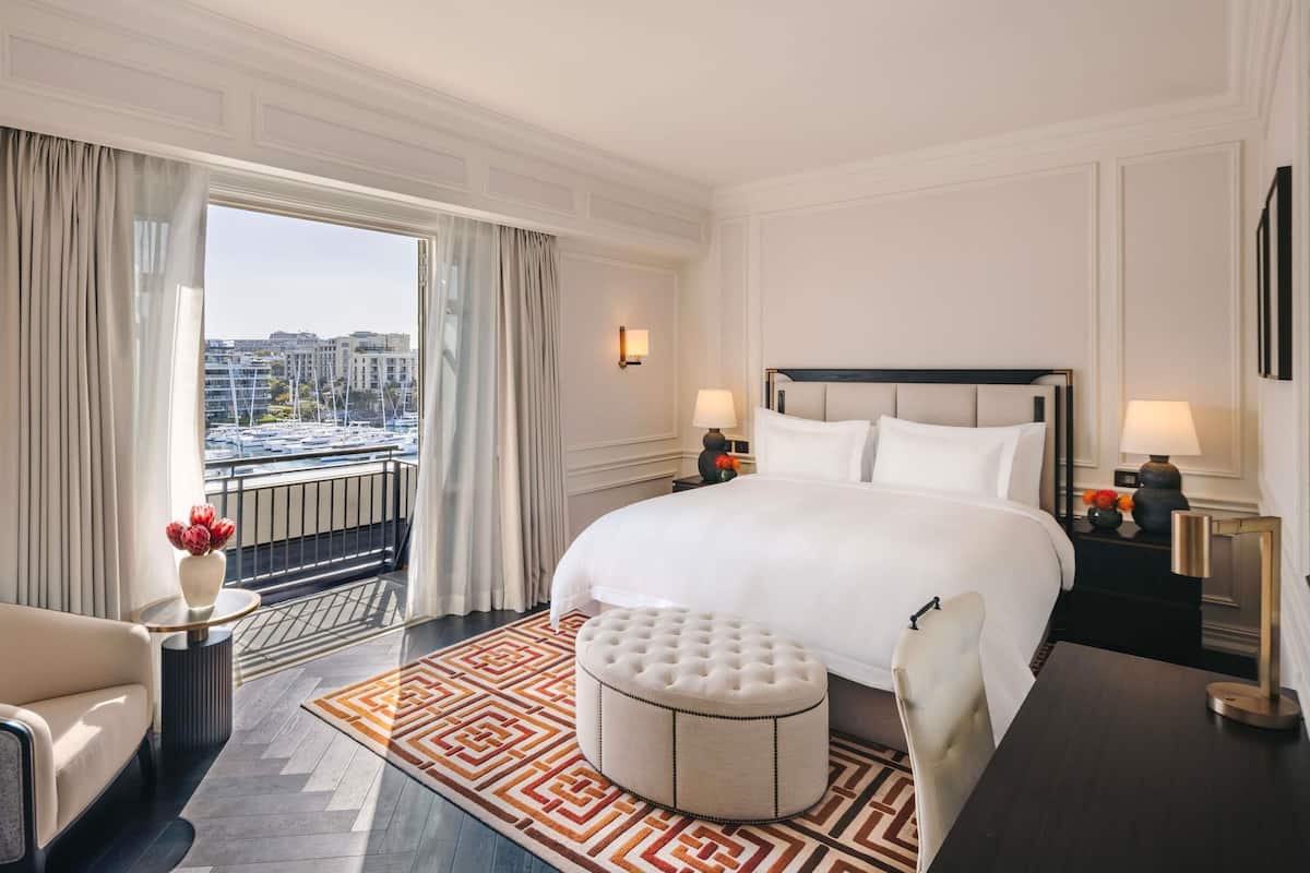 Habitación de hotel con una cama grande, un otoman redondo, una silla, una mesita y vistas a la ciudad por la ventana.