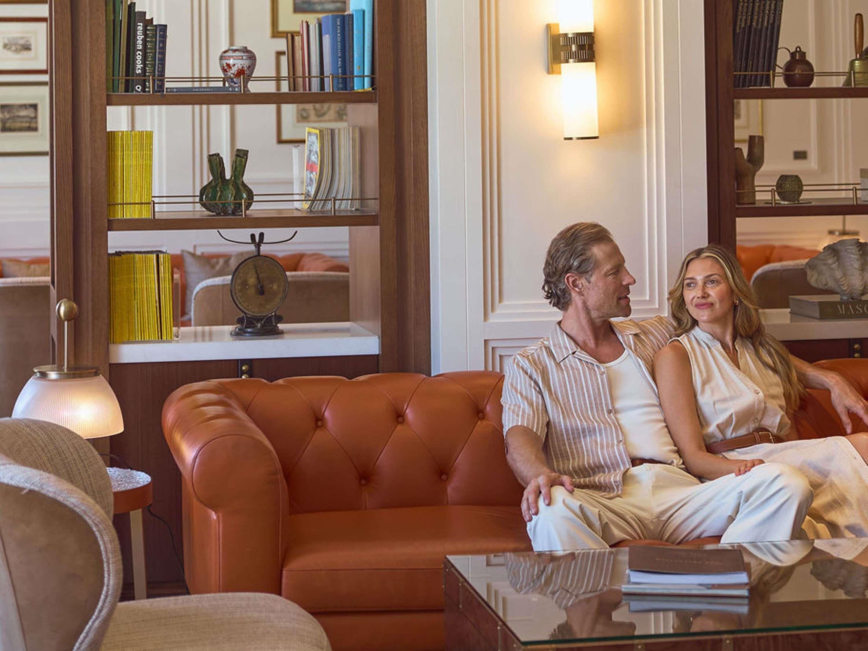 A living room with a brown leather sofa, a glass coffee table, and a bookshelf filled with books and decorative items.