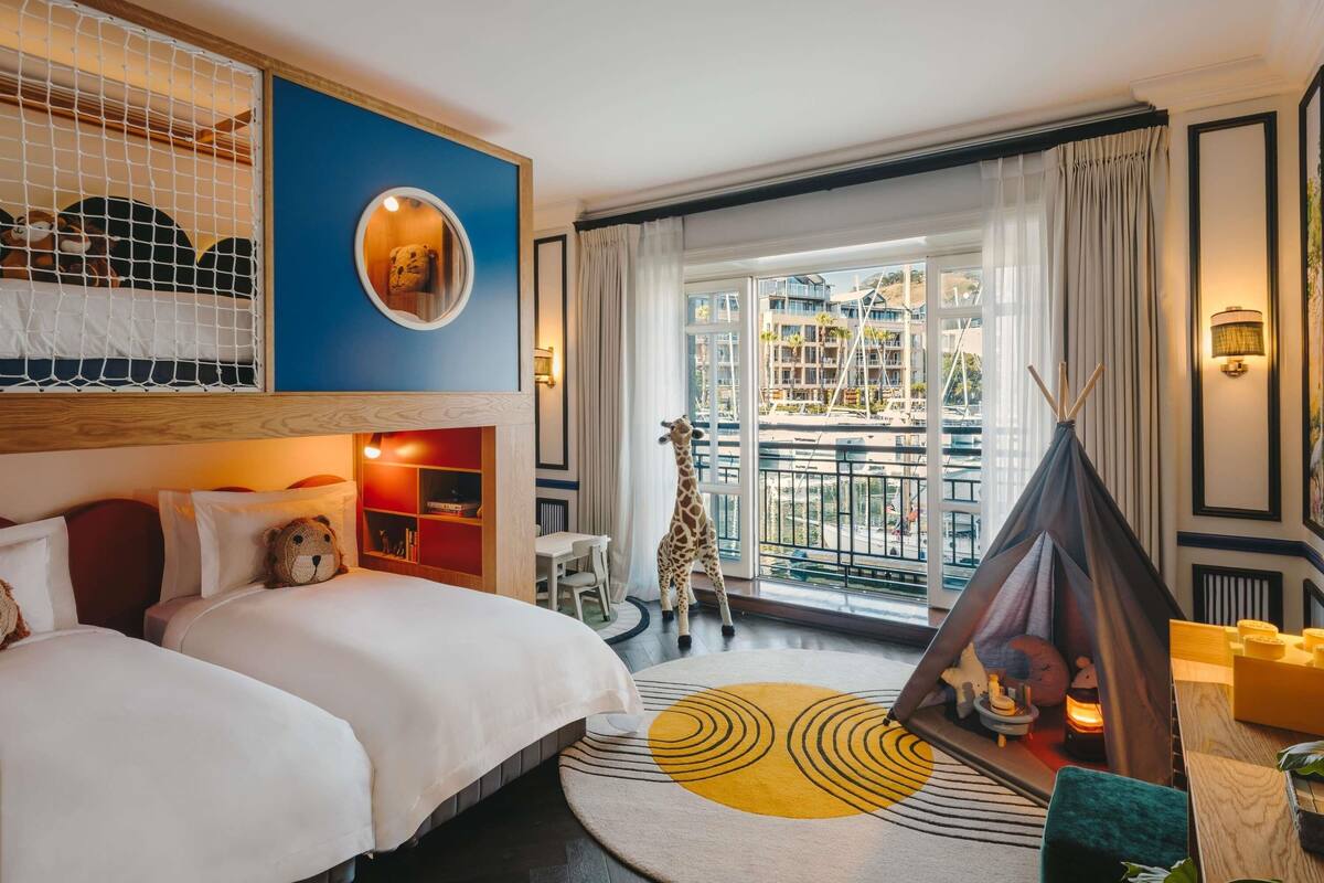 Una habitación de hotel con cama, una mesita, una silla, una ventana con vistas a edificios, una tienda de campaña y una lámpara.