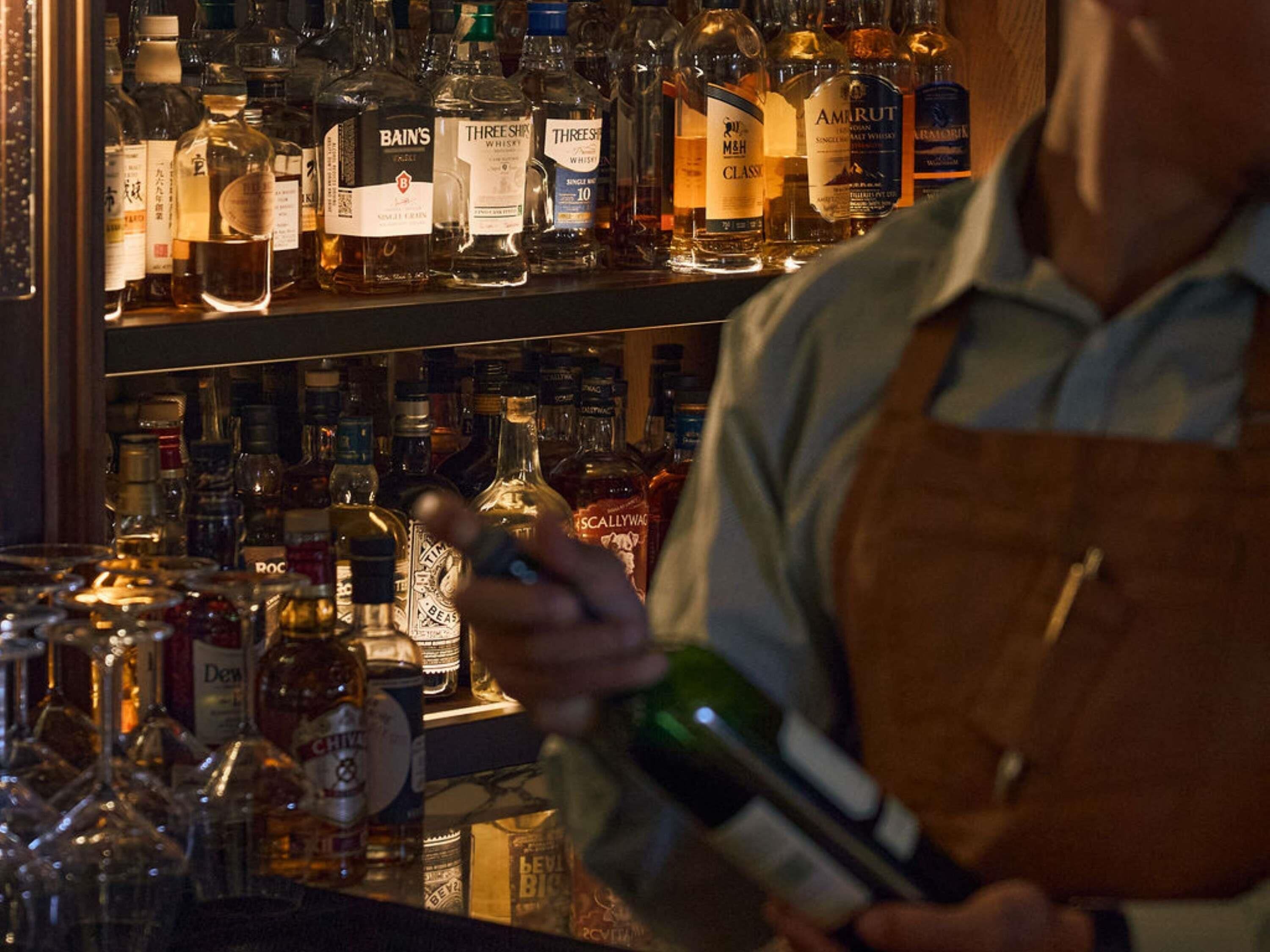 Un cantinero sosteniendo una botella, con estantes llenos de diferentes botellas de licor detrás.