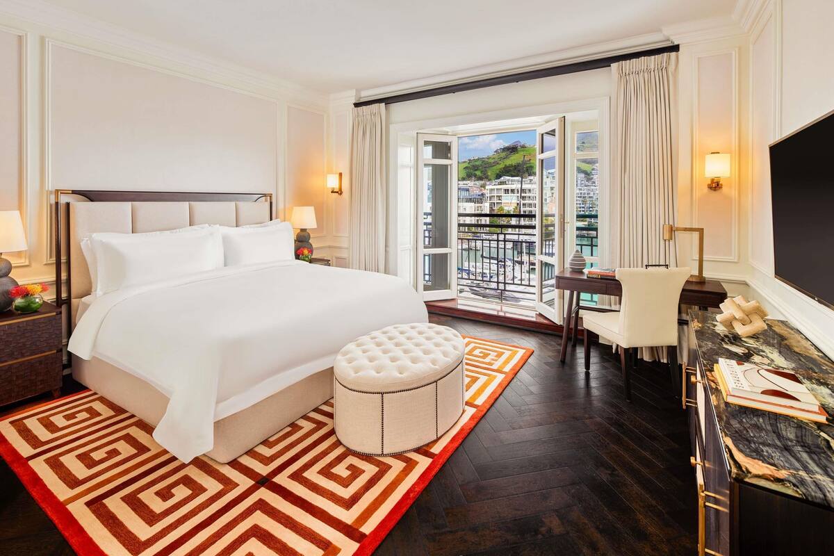 Habitación de hotel con una cama grande, un reposapiés pequeño y redondo, un escritorio con una silla y vistas a un paisaje urbano desde un balcón.